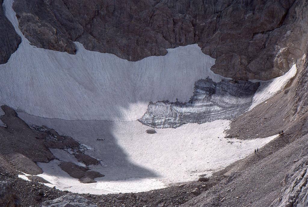El hielo glaciar, de color azul y estratificado, adosado a la pared suroeste del Jou Negro aflora parcialmente bajo la nieve y la neviza. La nieve marca, parcialmente, uno de los canales de avalancha. Los montículos que sobresalen por encima de la neviza están formados por clastos congelados. Varias personas, situadas sobre la parte occidental de la morrena, sirven de escala. Fecha: Septiembre 1992. Autor: J.J. González Suárez.