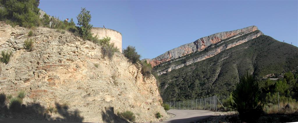 (679.063, 4.392.935; ETRS89 y huso 30). Afloramiento de la Fm Loriguilla en el corte de la carretera CV-3960 de acceso a la presa del embalse de Loriguilla. Al fondo, la ladera del otro lado del río Turia, mostrando esta misma formación con el característico relieve alomado con escaso desarrollo de suelo edáfico y vegetación de matorral mediterráneo, y encima el relieve estructural y crestón de los Calderones, originado por erosión diferencial en la Fm Higueruelas.