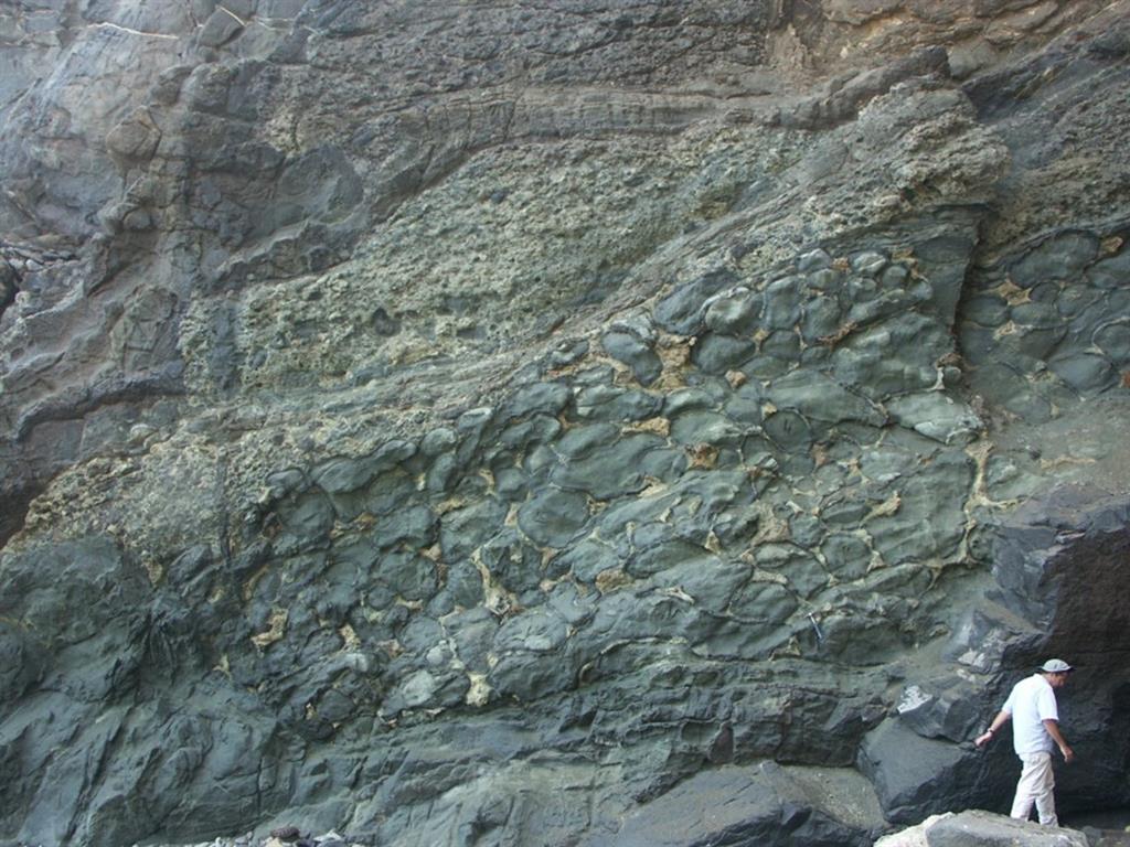 Afloramiento de lavas almohadilladas con brechas de fragmentos de almohadillas asociadas (Formación de Basaltos de El Valle, Grupo Volcánico Submarino), en la parte baja. En la parte alta aparecen las traquitas de la Caleta del Barco (Grupo Volcánico de Transicón). Playa de Los Mozos